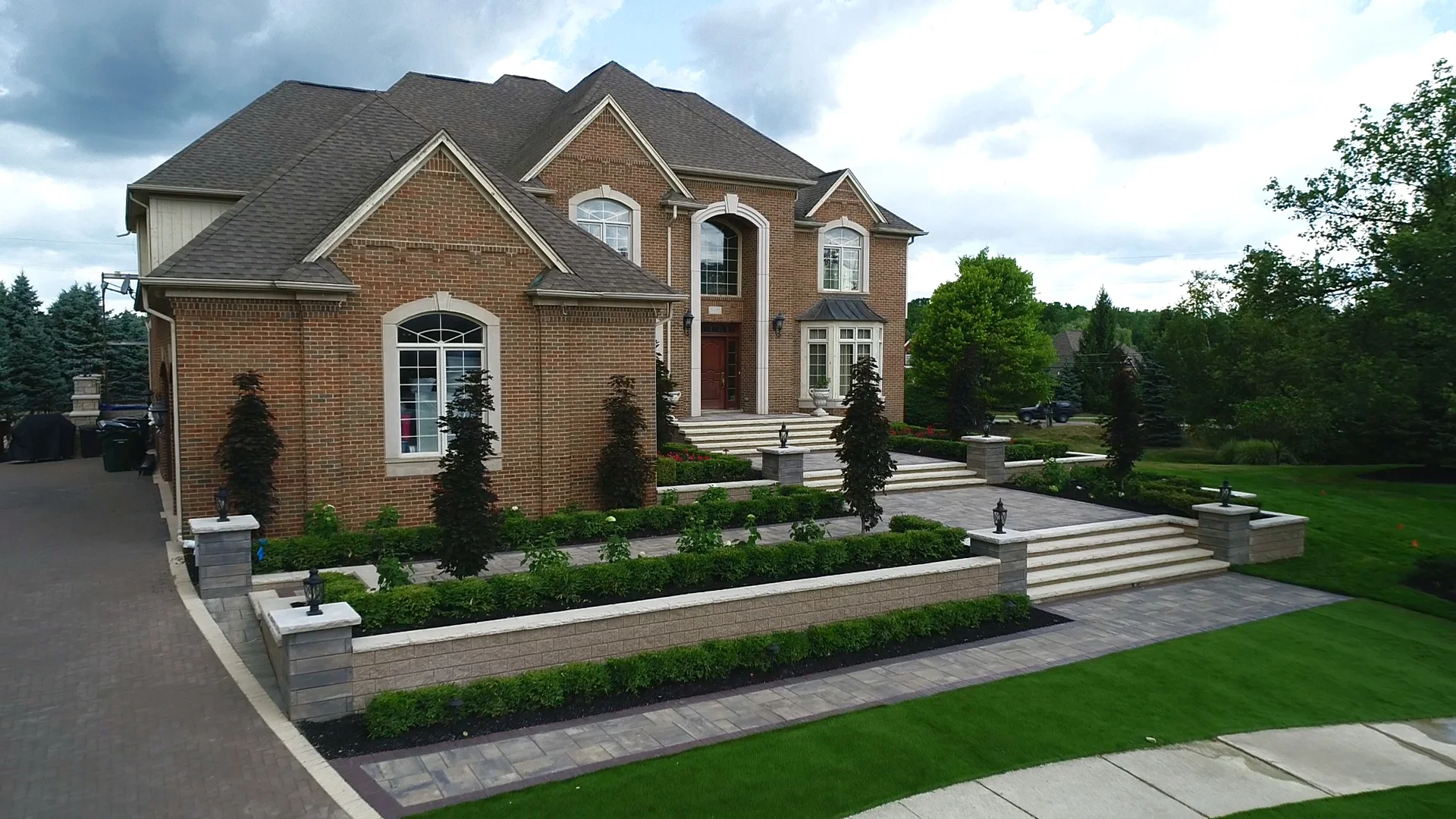 A large brick house featuring tiered landscaping with stone retaining walls, manicured shrubs, and a paved walkway leading to the front entrance.