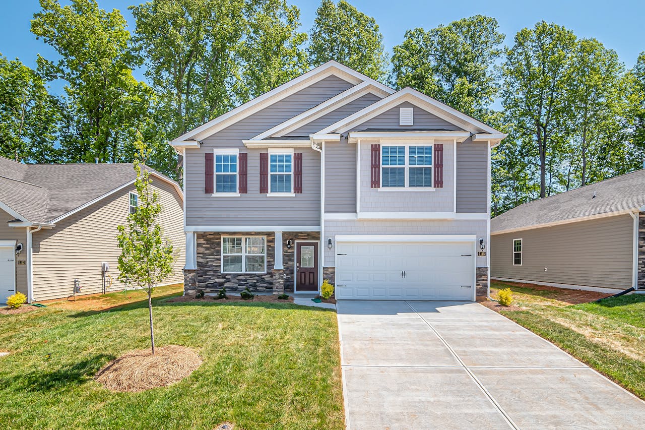 A two-story suburban home with gray siding, stone accents, dark red window shutters, and an attached two-car garage, featuring a concrete driveway and a front lawn.