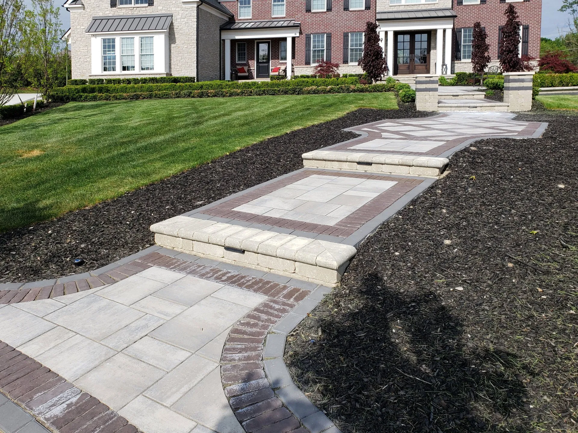 A paved stone walkway with steps leading to the front entrance of a brick suburban home, surrounded by mulch and a manicured lawn.